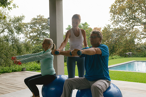 Side view of Caucasian female trainer assisting active senior couple to exercise with dumbbell in the porch at home
