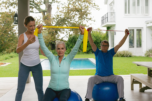 Front view of Caucasian female trainer assisting active senior couple to exercise with resistance band in the porch at home