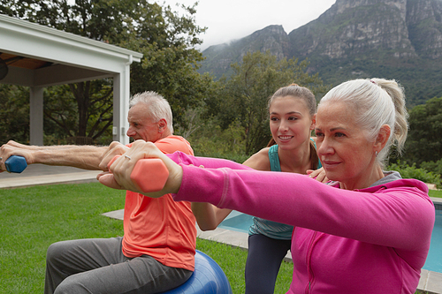 Side view of Caucasian female trainer assisting active senior Caucasian couple to exercise with dumbbells in the backyard