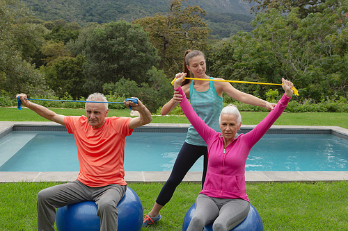 Front view of Caucasian female trainer assisting active senior Caucasian couple to exercise with resistance band in the backyard
