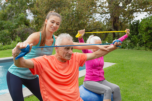 Side view of Caucasian female trainer assisting active senior Caucasian couple to exercise with resistance band in the backyard
