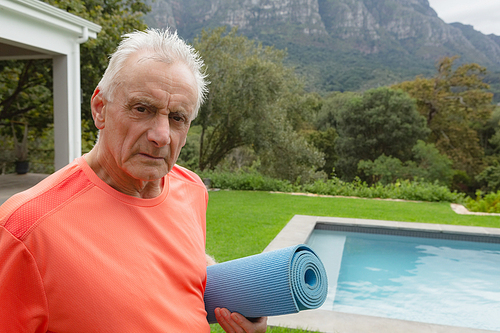Portrait of active senior Caucasian man standing with exercise mat near poolside in the backyard