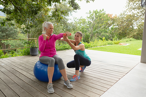 Front view of beautiful Caucasian female trainer assisting active senior Caucasian woman to exercise with dumbbells in the porch