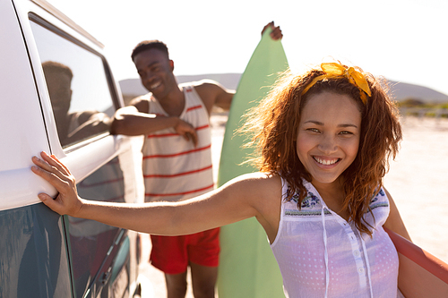 Front view of happy young Mixed-race couple with surfboard leaning on camper van at beach