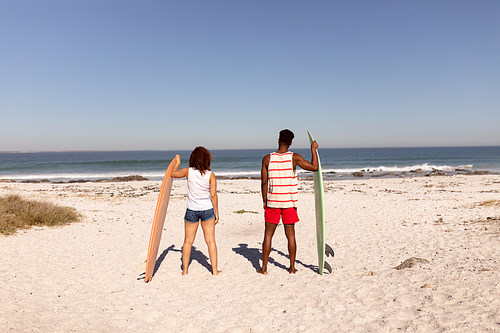 Rear view of couple standing with surfboard on beach in the sunshine