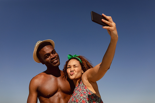 Low angle view of happy young Mixed-race couple taking selfie with mobile phone on beach in the sunshine