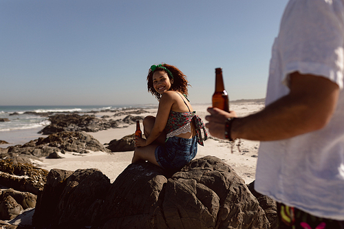 Rear view of young Mixed-race woman with beer bottle sitting on rock and looking at man on beach in the sunshine