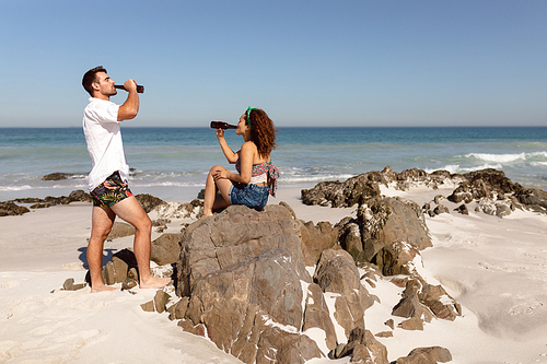 Side view of young Mixed-race couple drinking beer on beach in the sunshine