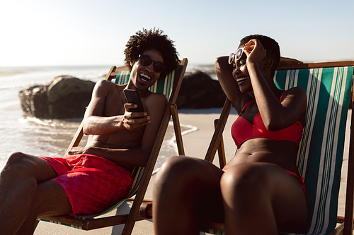 Front view of happy African-american couple having fun on mobile phone while relaxing in a beach chair on the beach