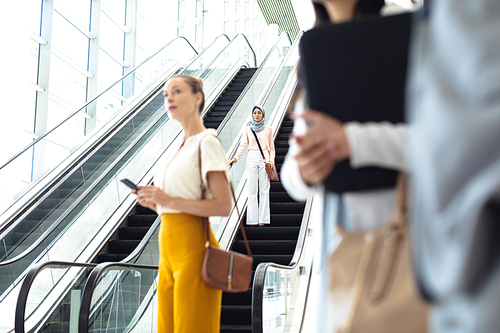 Front view of young female executive taking escalator while Caucasian female executive look straight ahead  in modern office