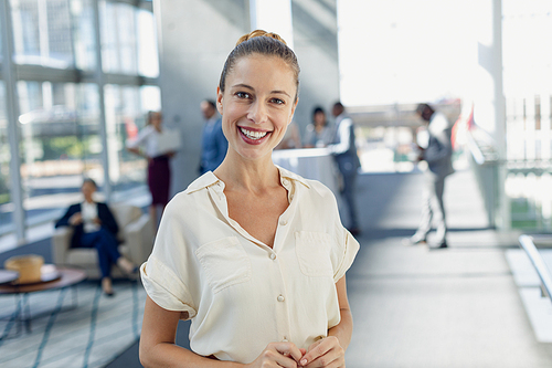 Front view of Caucasian female executive  while standing in modern office. Behind her, colleagues interacting with each other.