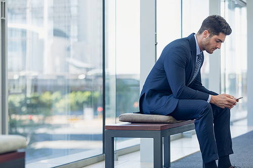 Side view of young Caucasian businessman looking at mobile phone while sitting on bench in modern office.