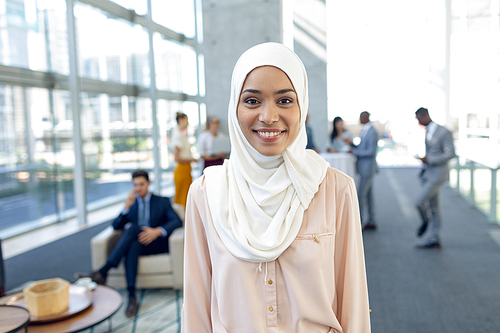 Front view of young pretty businesswoman in hijab  while standing in modern office. Behind her, colleagues interacting with each other.