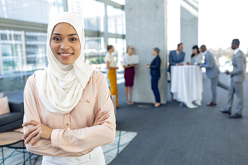 Front view of young pretty businesswoman in hijab  while standing in modern office.  Behind her, colleagues interacting with each other.