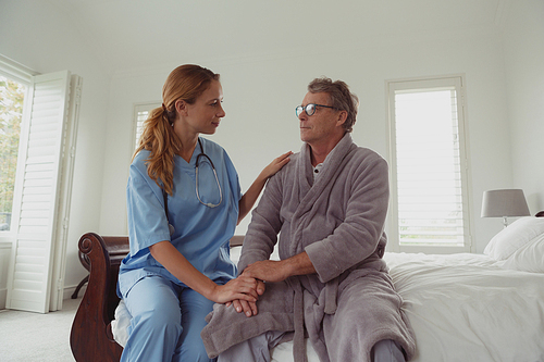 Front view of beautiful Caucasian female doctor consoling active senior man on bed in bedroom at comfortable home