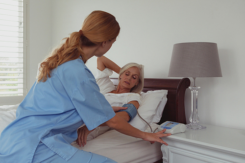 Rear view of Caucasian female doctor checking blood pressure of active senior woman in bed in bedroom at home