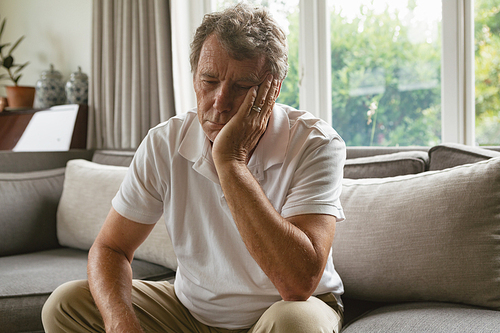 Front view of sad active senior Caucasian man with hand on face sitting on sofa in a comfortable home