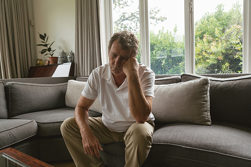 Front view of sad active senior Caucasian man with hand on face sitting on sofa in a comfortable home