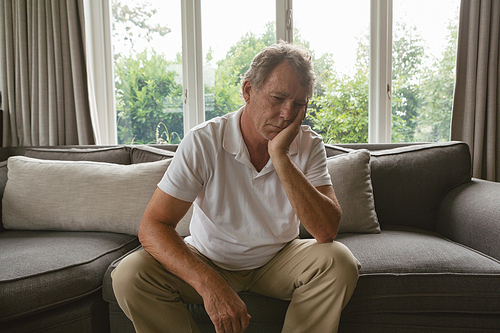 Front view of sad active senior Caucasian man with hand on face sitting on sofa in a comfortable home
