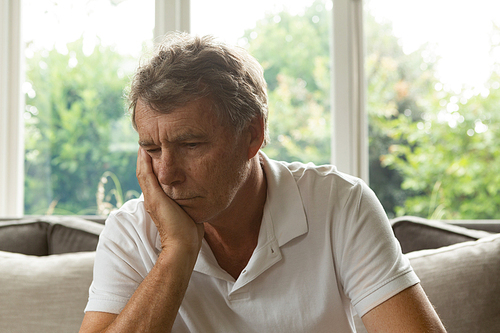 Front view of sad active senior Caucasian man with hand on face sitting on sofa in a comfortable home