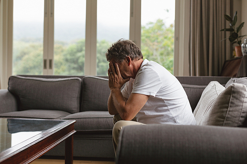 Side view of sad active senior Caucasian man covering his face with hands while sitting on sofa in a comfortable home