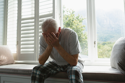 Front view of active senior Caucasian man covering face with hands on window seat in bedroom at home