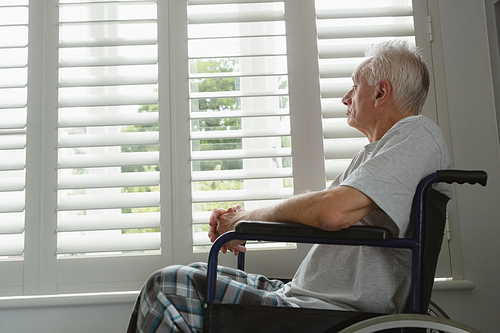 Side view of thoughtful active senior Caucasian man in wheelchair looking through window at home