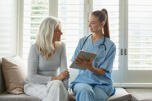 Front view of active senior Caucasian woman and female doctor using digital tablet on window seat at home