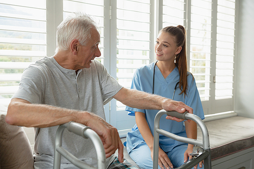 Side view of Caucasian female doctor interacting with active senior man on window seat at home