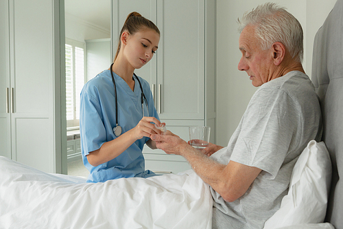 Side view of Caucasian female doctor giving medicine to active senior Caucasian male patient in bedroom at home