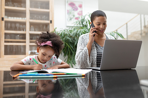 Front view of African american girl doing her homework while mother using laptop and talking on mobile phone at home