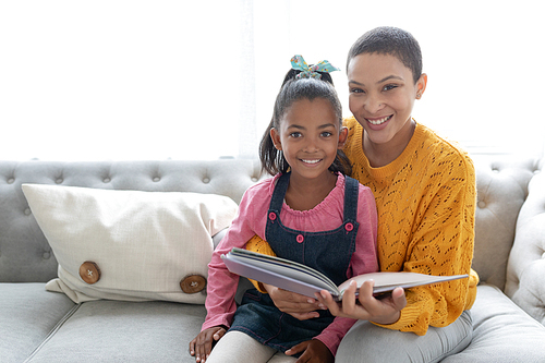 Portrait of African american mother and daughter reading a story book on a sofa in living room at home