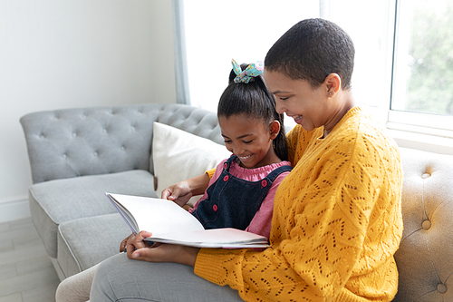 Side view of African american mother and daughter reading a story book on a sofa in living room at home