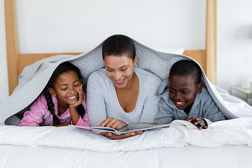 Front view of African american Mother and children reading a story book while relaxing under a blanket in bedroom at home
