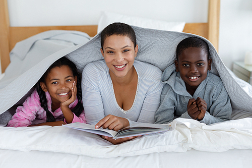 Front view of African american mother and children reading a story book while relaxing under a blanket in bedroom at home