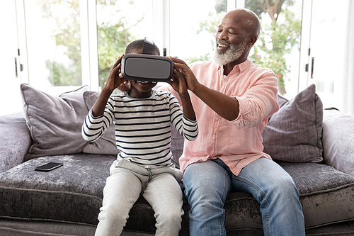 Front view of African american Grandfather helping his grandson to wear virtual reality headset in living room at home