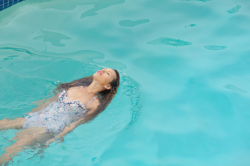 High angle view of beautiful Caucasian woman with eyes closed floating in swimming pool at the backyard of homeoman with eyes closed floating in swimming pool in the backyard at home. Summer fun at home by the swimming pool