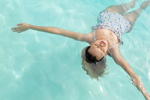 High ange view of beautiful Caucasian woman with eyes closed floating in swimming pool at the backyard of home. Summer fun at home by the swimming pool