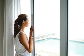 Side view of thoughtful mixed-race woman looking through window in living room at home. Summer fun at home by the swimming pool