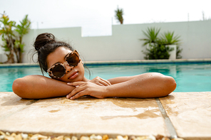 Front view of beautiful mixed-race woman in sunglasses standing at the edge of swimming pool in the backyard at home. Summer fun at home by the swimming pool