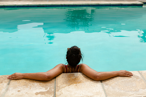 Rear view of mixed-race woman relaxing at the edge of swimming pool in the backyard at home. Summer fun at home by the swimming pool