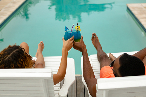High angle view of diverse couple toasting glasses of cocktail while relaxing on a sun lounger near swimming pool at the backyard of home. Summer fun at home by the swimming pool