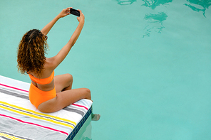 High angle view of beautiful mixed-race woman taking selfie with mobile phone while sitting at the edge of swimming pool. Summer fun at home by the swimming pool