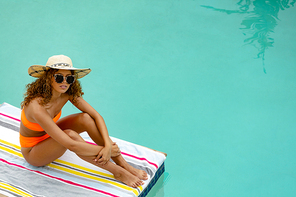 High angle view of beautiful mixed-race woman in sunglasses and hat sitting at the edge of swimming pool. Summer fun at home by the swimming pool