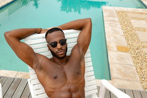 High angle view of handsome shirtless African-american man in sunglasses relaxing on a sun lounger near swimming pool. Summer fun at home by the swimming pool