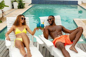 Front view of happy diverse couple toasting glasses of cocktail while relaxing on a sun lounger near swimming pool. Summer fun at home by the swimming pool