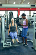 Front view of pretty young African-american female trainer assisting active senior Caucasian woman to work out on leg curl machine in fitness center. Bright modern gym with fit healthy people working out and training