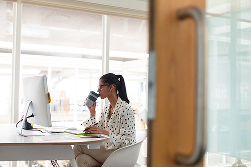 Front view of beautiful mixed-race female graphic designer having coffee while using graphic tablet at desk in office. Modern casual creative business concept