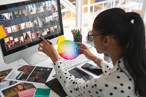 High angle view of pretty mixed-race female graphic designer working on computer and holding colour swatch at desk in office. Modern casual creative business concept