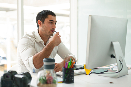 Front view of thoughtful young Caucasian male graphic designer looking at computer at desk in office. Modern casual creative business concept